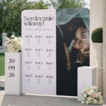 Fotografia w orientacji pionowej prezentuje ściankę z planem stołów weselnych w eleganckiej, jasnej aranżacji plenerowej. Projekt łączy czytelną listę gości z estetycznym układem graficznym i personalizowanym motywem ślubnym.