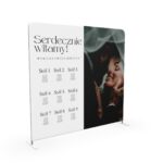 Pionowa ścianka weselna z planem stołów i zdjęciem pary młodej w eleganckiej aranżacji plenerowej.