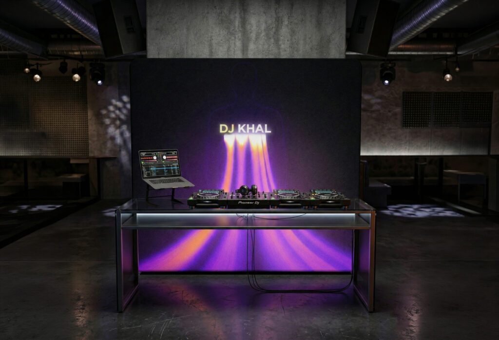 Scianka-eventowa-DJ-konsola-klub Stanowisko DJ-a z profesjonalną konsolą i laptopem, ustawione na tle nowoczesnej ścianki eventowej z neonowym napisem DJ KHAL i fioletowo-pomarańczowymi promieniami światła w surowym wnętrzu klubu.